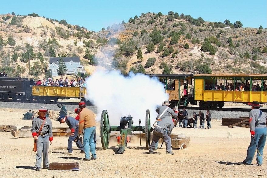 A historical reenactment: soldiers fire a large black cannon as smoke billows, with a yellow passenger train and spectators in the background.
